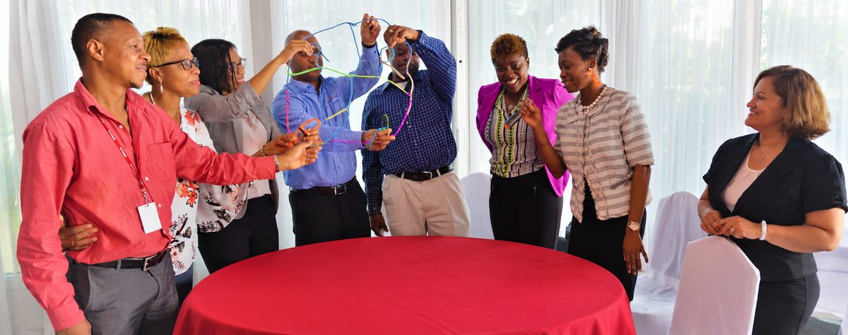 A workshop group standing around a table, passing a web of
                    coloured string between them, mid-exercise.