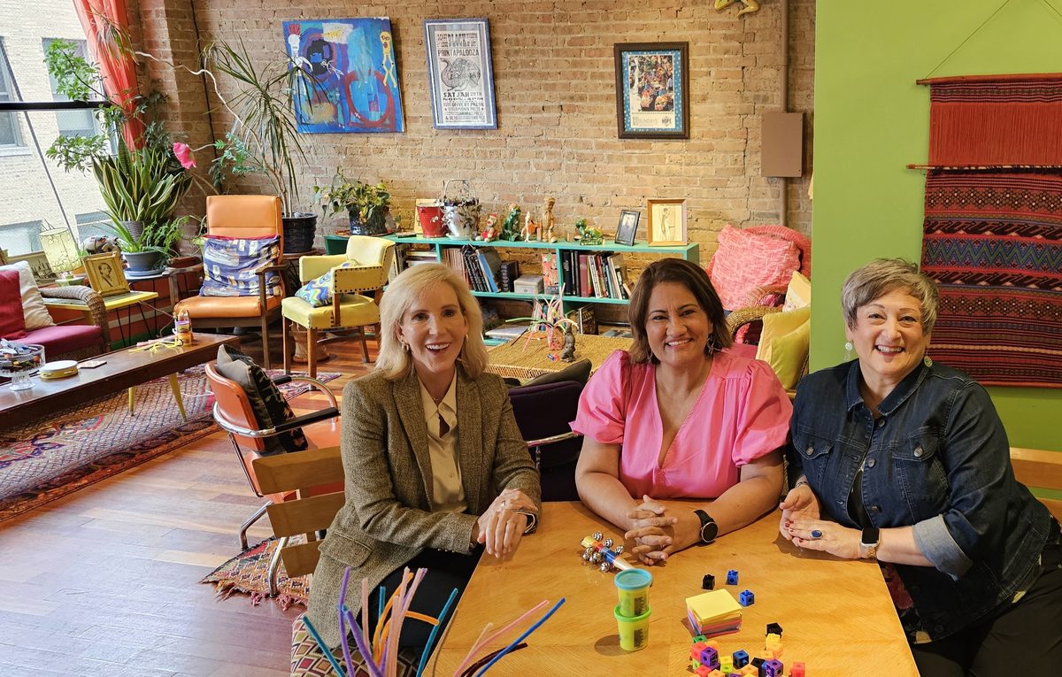 Three colleagues seated around a workshop table with
                      building blocks and materials, talking and smiling.